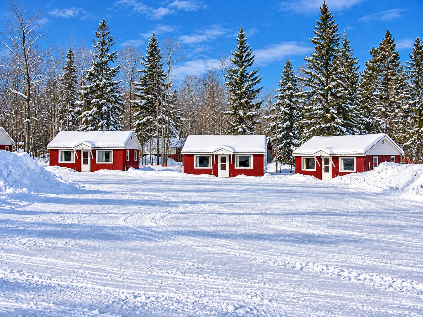 Row of red cabins with snow-covered roofs and groomed snow between them on a clear winter day