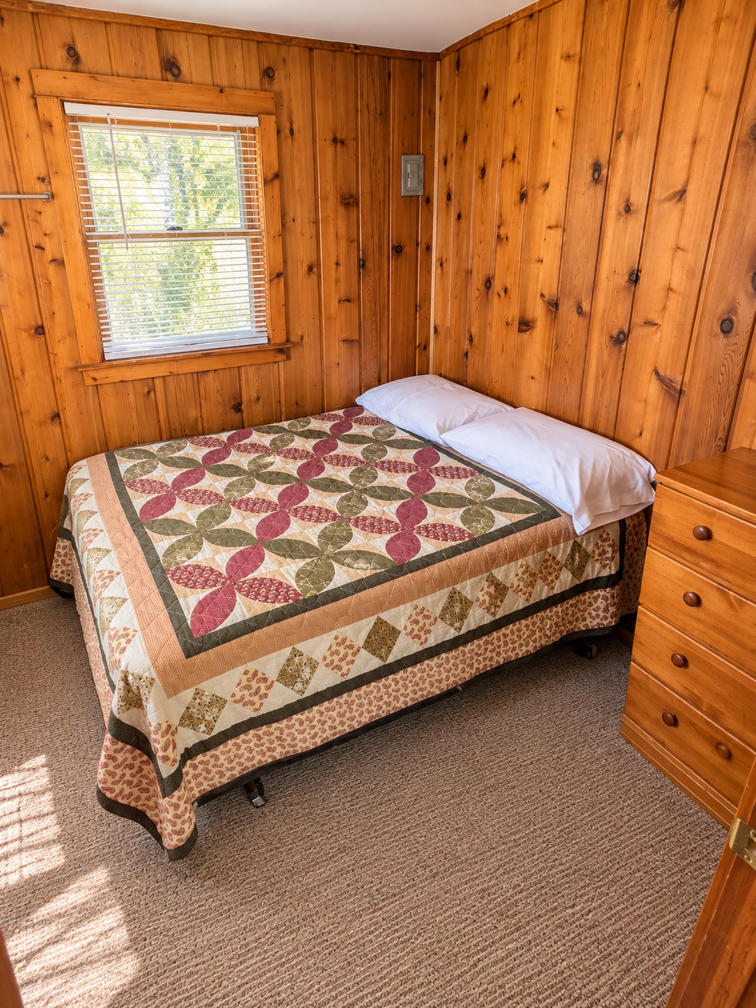 Cabin bedroom with a quilt-covered double bed, pine dresser, and window looking into the trees