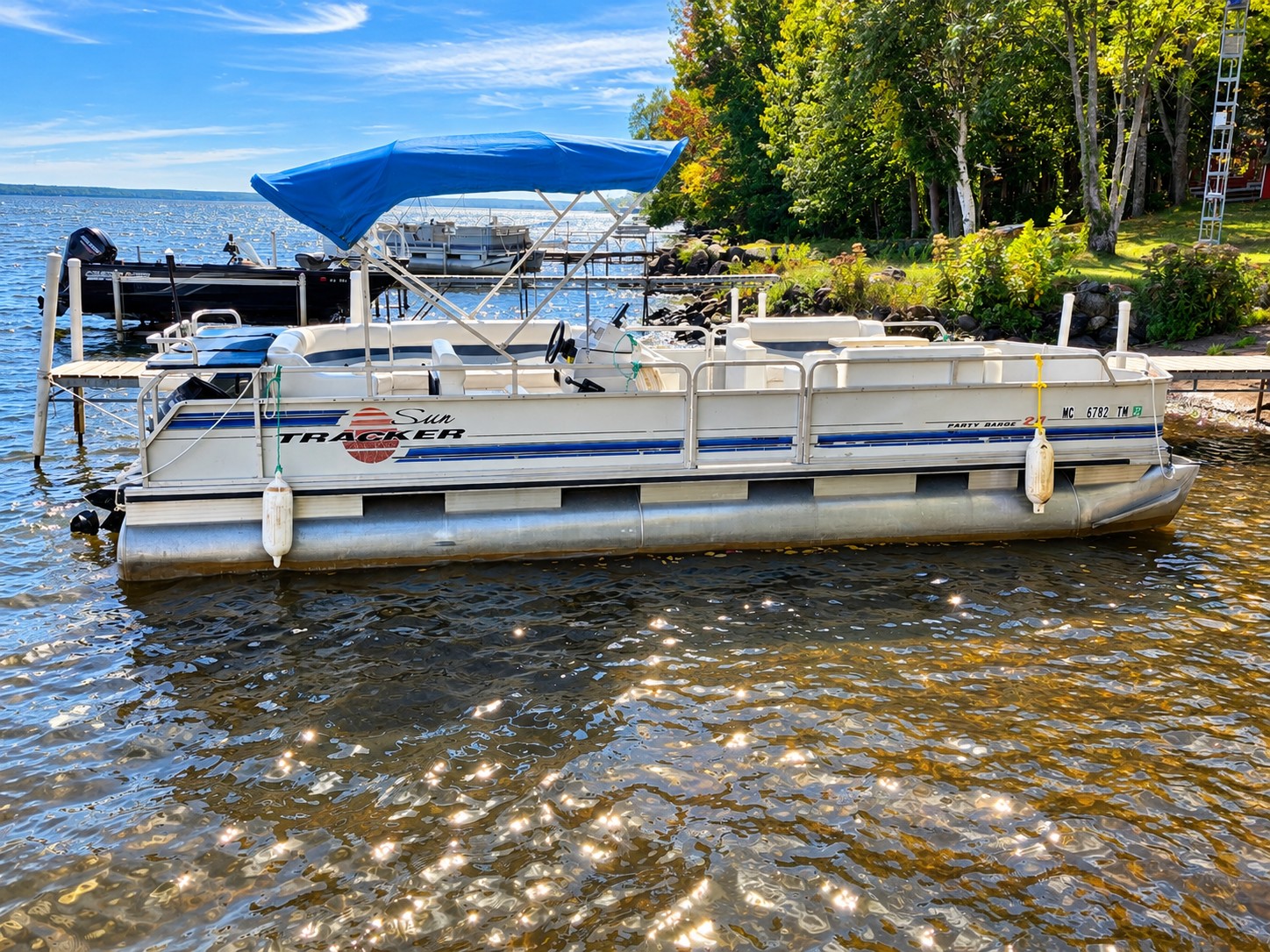 Sun Tracker Party Barge pontoon with a blue canopy moored along the shoreline in early fall